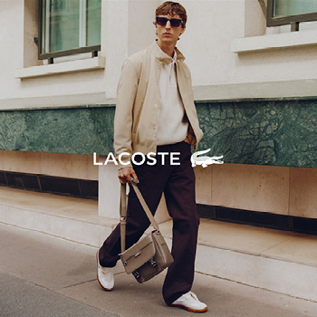 Man wearing beige jacket, white jumper, dark trousers and white trainers, carrying grey leather bag, Lacoste logo overlay.