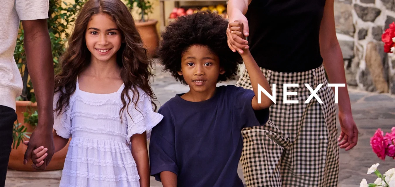 Una famiglia che cammina lungo una strada in acciottolato. L'uomo indossa un maglione leggero e pantaloni corti; i bambini hanno indumenti con motivi vari. Sono presenti piante fiorite.