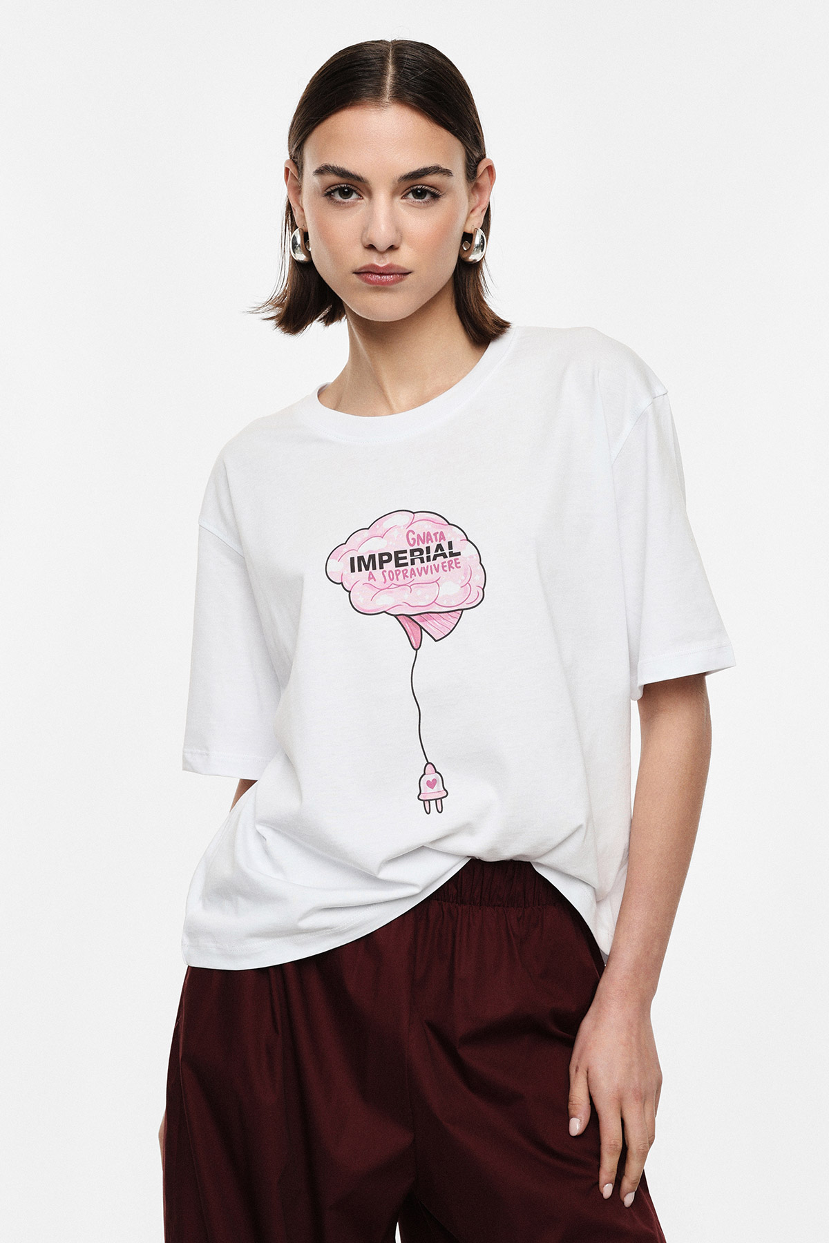 Model in white oversized graphic T-shirt featuring pink brain balloon print and burgundy trousers, studio fashion portrait