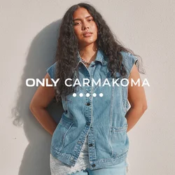 Donna con capelli lunghi e ricci che indossa un gilet di jeans senza maniche e pantaloni azzurri, in piedi contro un muro semplice di colore chiaro.