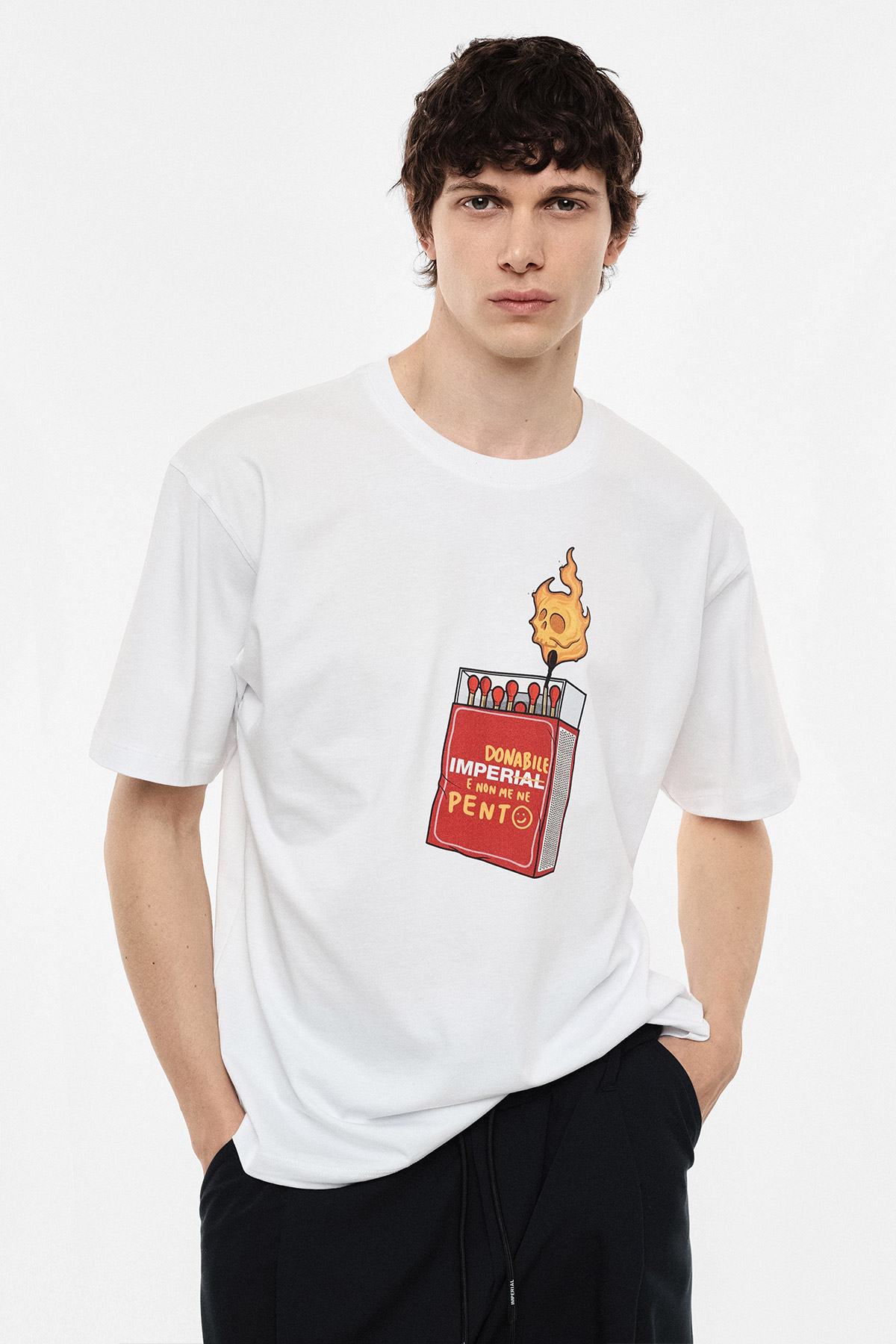 Male model wearing white T-shirt with printed red matchbox and flaming match graphic, paired with black trousers