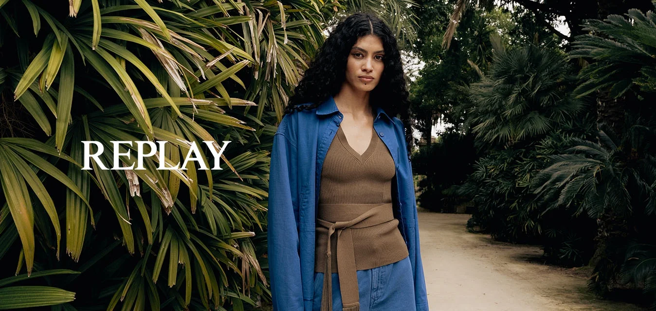 Donna con capelli ricci che indossa una giacca blu e pantaloni ampi, in piedi su un sentiero di terra circondato da piante tropicali dense e palme.