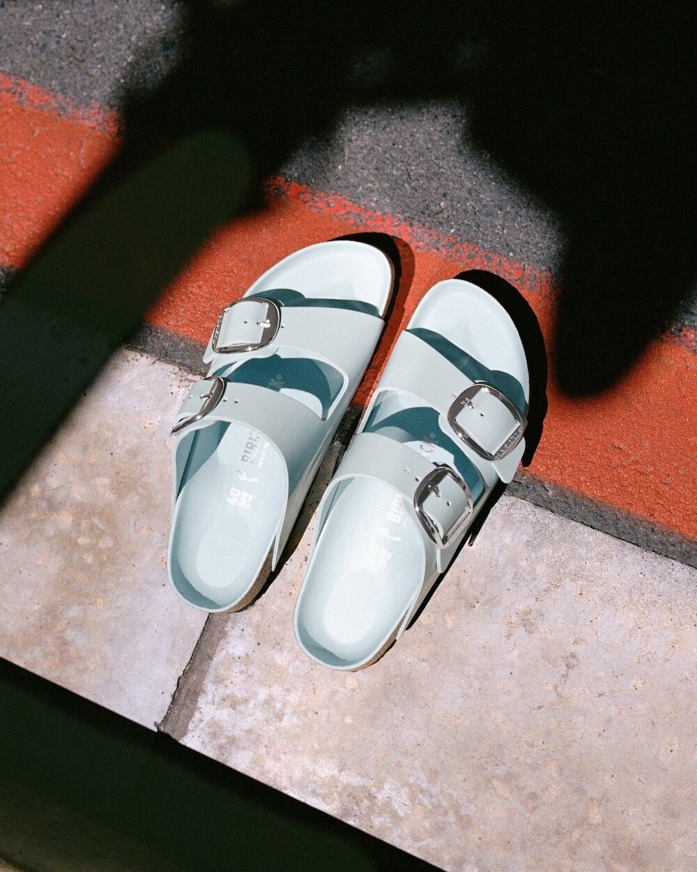 Pair of mint-green double-buckle sandals on tiled pavement beside a red-painted curb, lit by sunlight with shadows