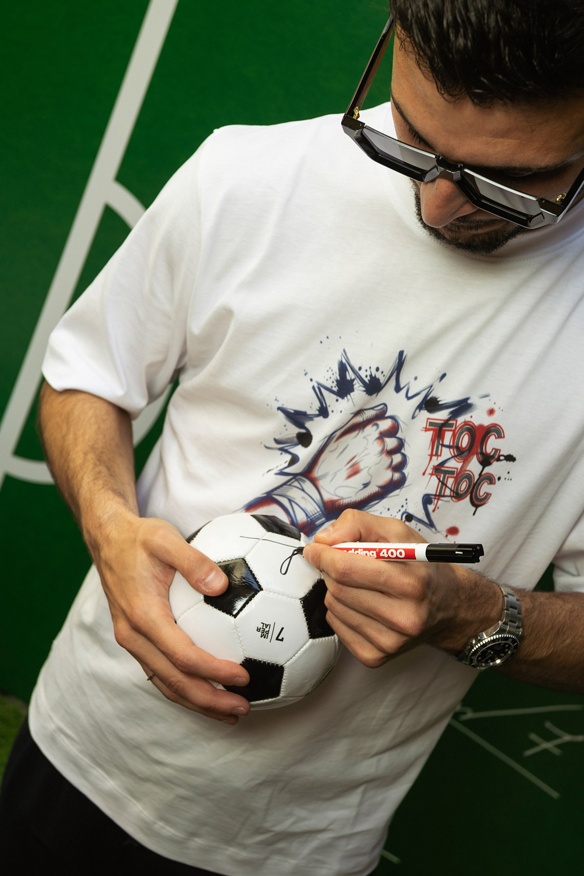 Man signing a football with a black marker while wearing sunglasses and a white graphic T-shirt, close-up on hands and ball