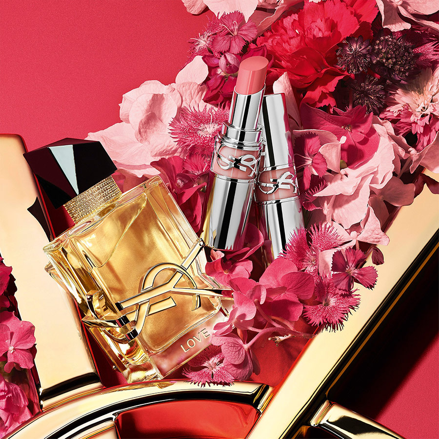 Profumo YSL Libre inciso "LOVE" e due rossetti rosa tra fiori vivaci.