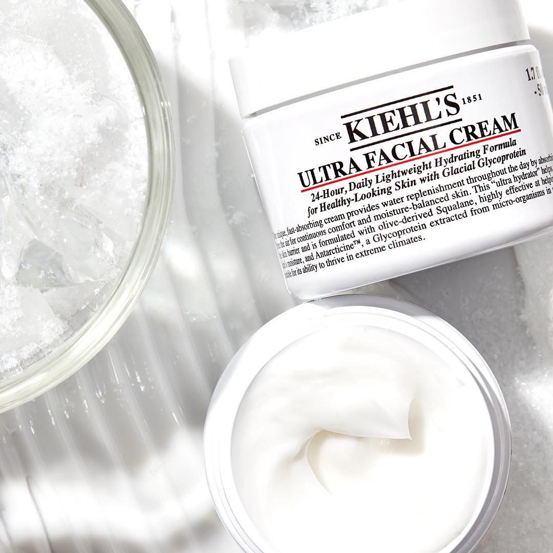 Kiehl's Ultra Facial Cream aperta accanto a una ciotola di ghiaccio su superficie bianca, con la texture cremosa in primo piano.