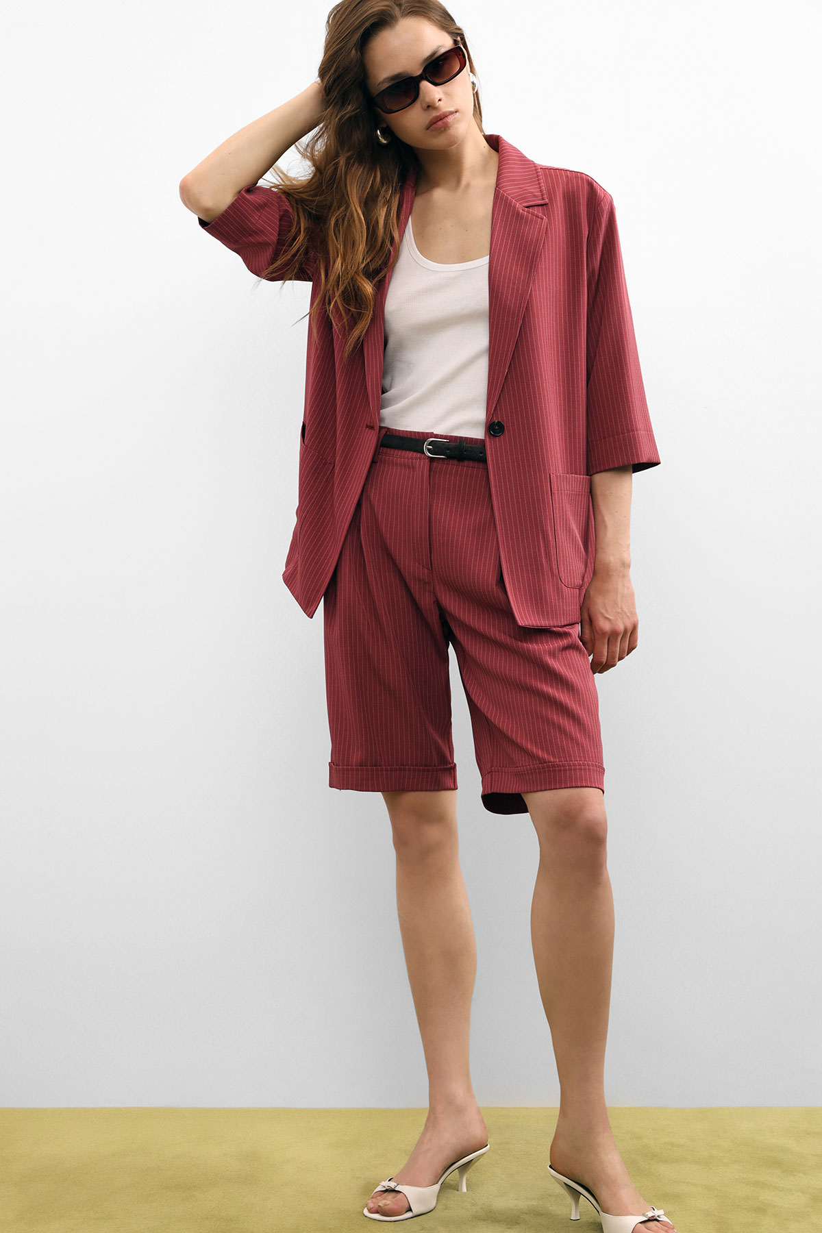 Model wearing red pinstripe oversized blazer and matching knee-length shorts, white tank top, black belt, sunglasses and cream heels