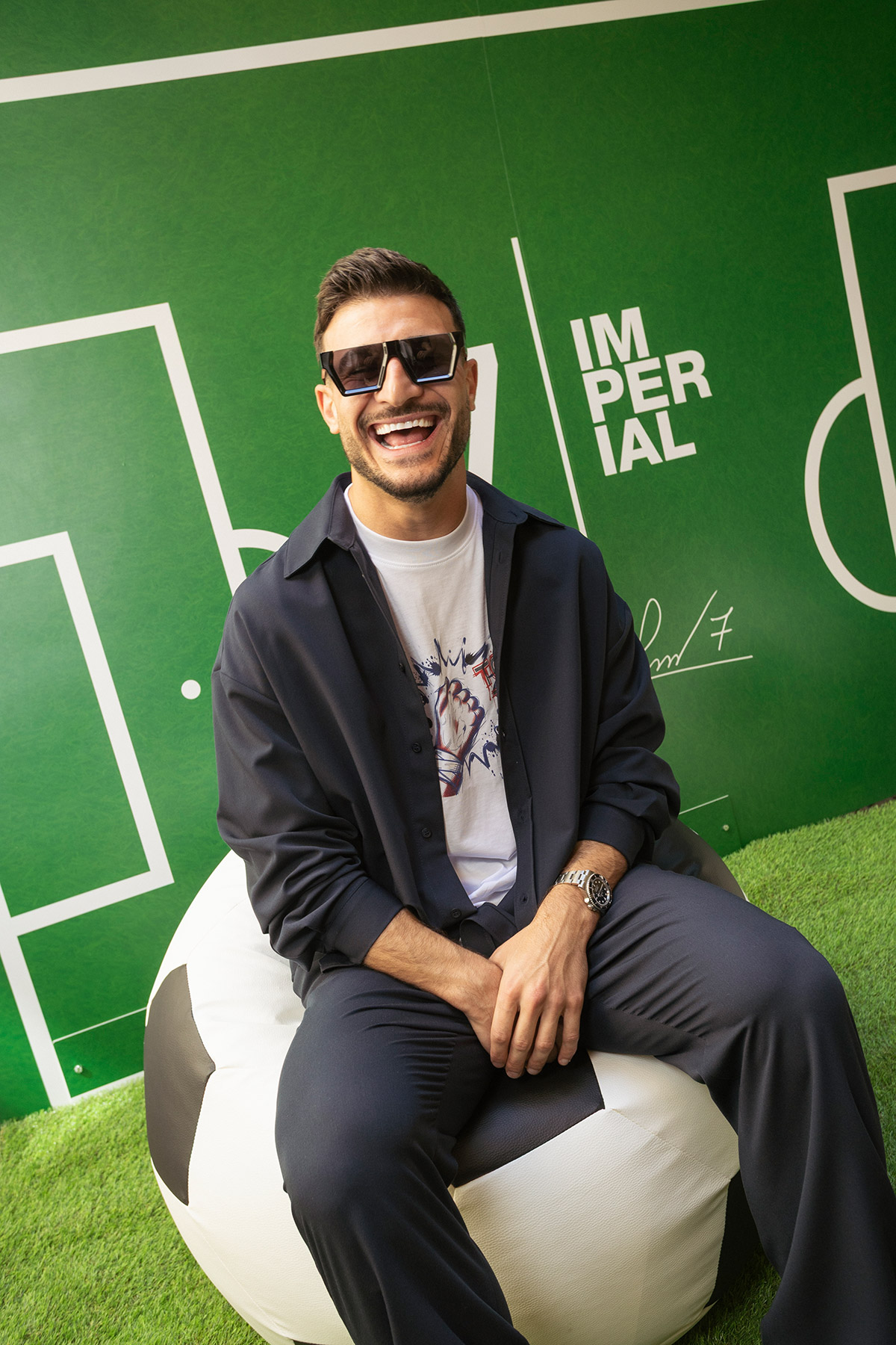 Smiling young man in sunglasses and casual navy outfit sitting on giant football beanbag against green pitch backdrop