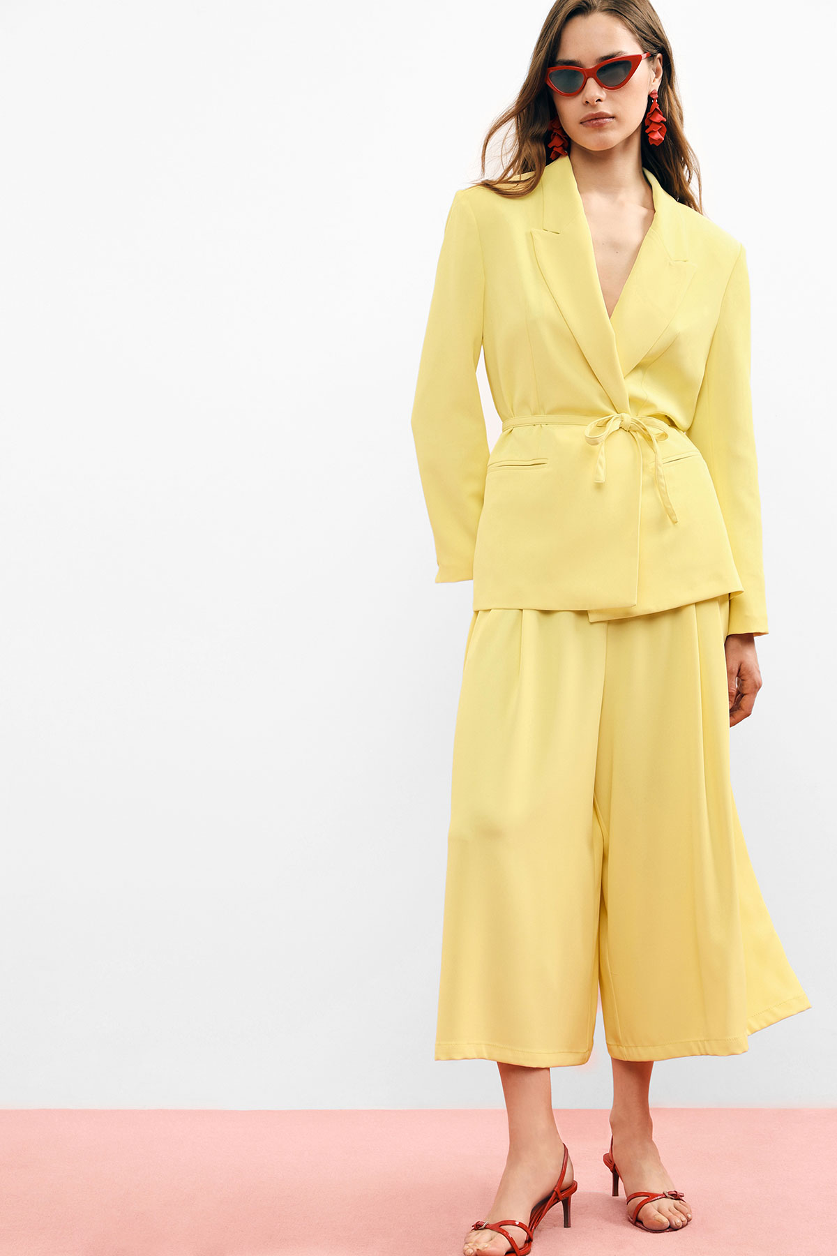 Woman in lemon-yellow belted blazer and wide trousers with red cat-eye sunglasses, red earrings and heels on pink floor