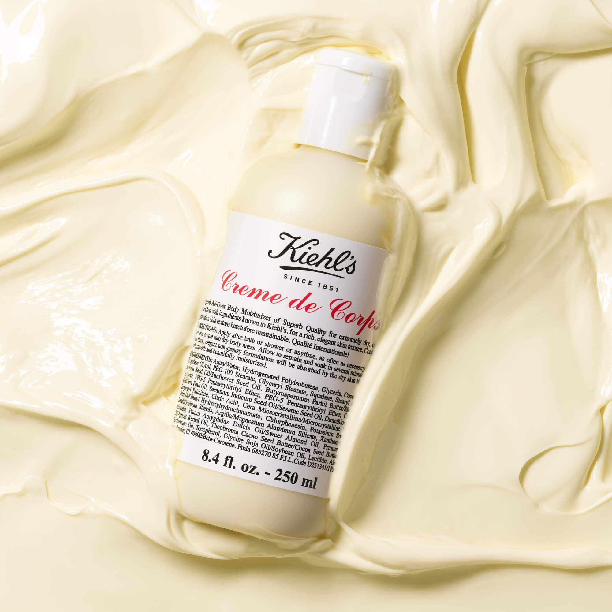 Flacone di Kiehl's Creme de Corps adagiato su un fondo di crema corpo color avorio, con texture ricca e vellutata.