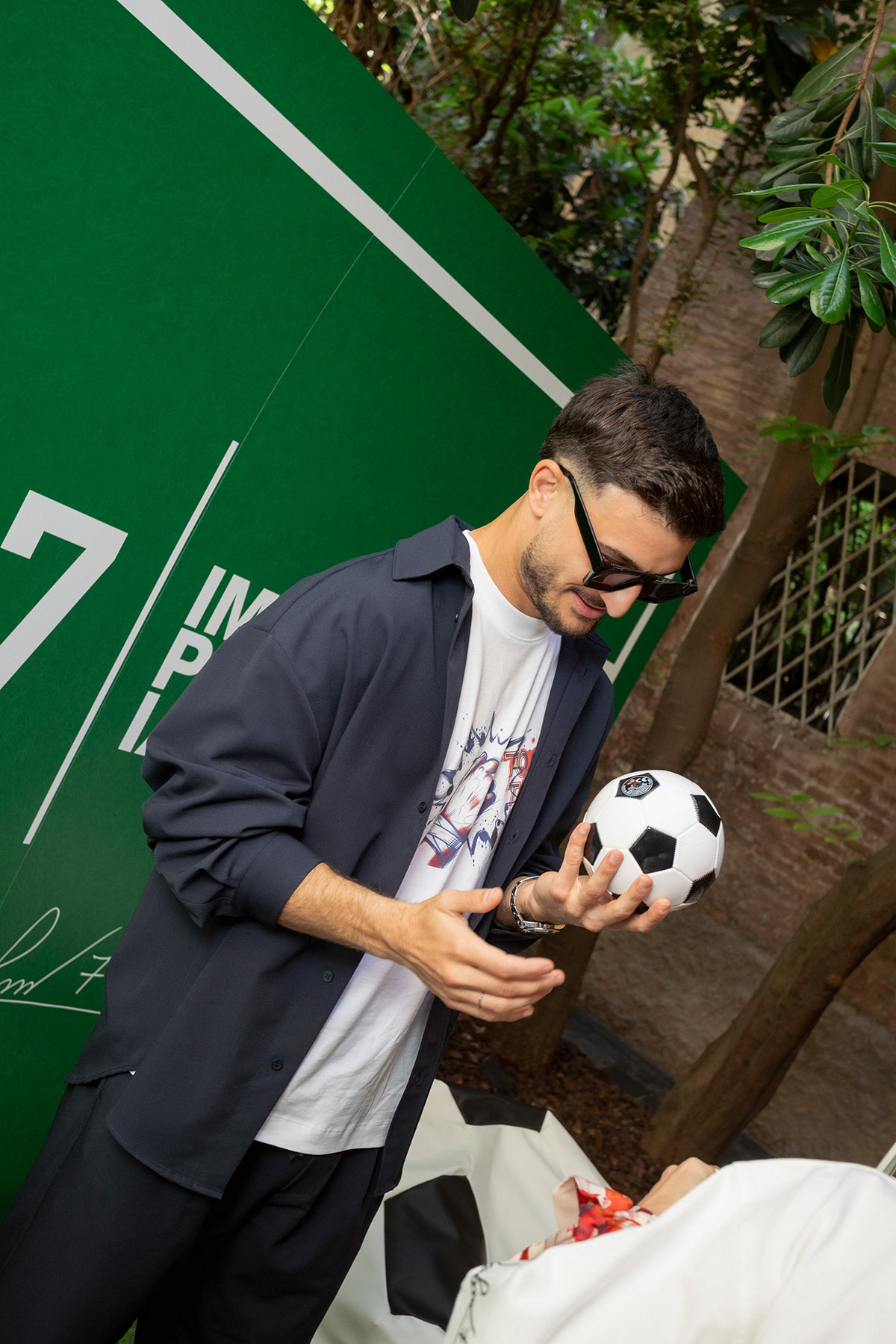 Young man wearing sunglasses and casual clothes holding a black-and-white football in front of a green promotional backdrop outdoors