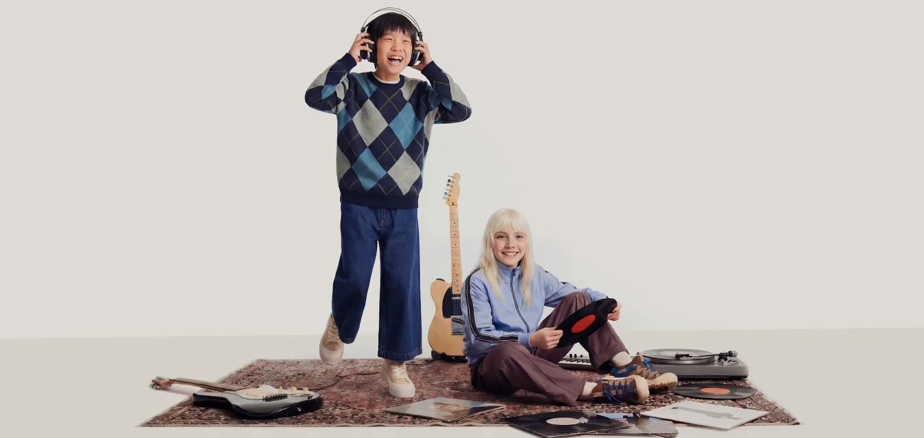 Due bambini con attrezzatura musicale: uno in piedi con cuffie, l'altro seduto su un tappeto che tiene un disco in vinile vicino a un giradischi e a delle chitarre.