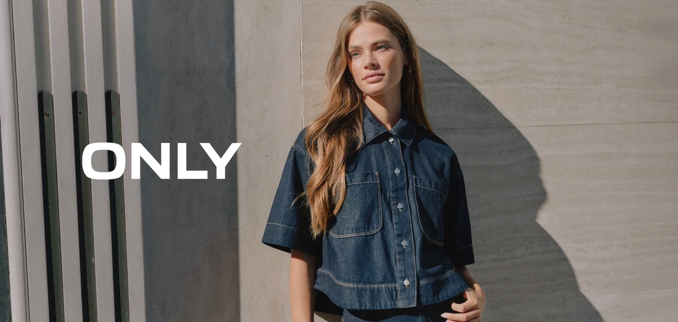 Giovane donna con capelli lunghi in piedi contro un muro di cemento, indossando una camicia a maniche corte in denim scuro e jeans a vita alta abbinati.