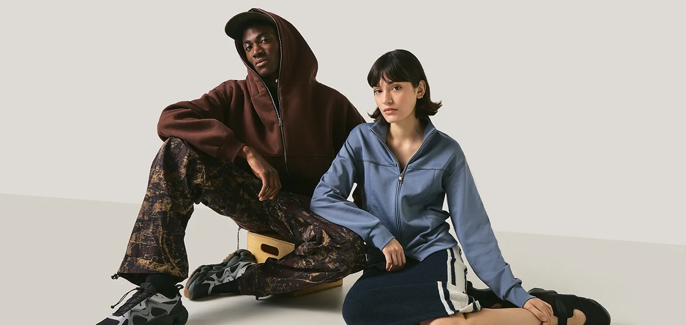 Due persone sedute su un pavimento semplice contro uno sfondo neutro, una indossa una felpa con cappuccio e pantaloni a fantasia, l'altra una giacca blu e una gonna.