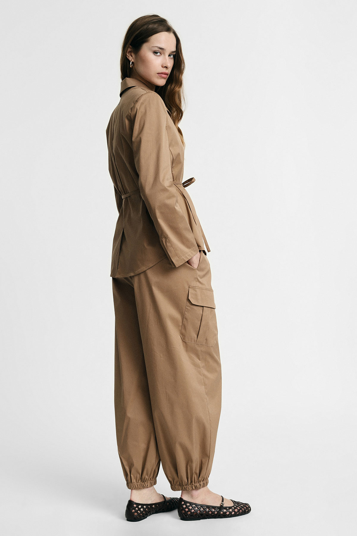 Woman wearing tan utility jacket and matching cargo trousers with elasticated ankles, standing side-on and looking back in black woven flats