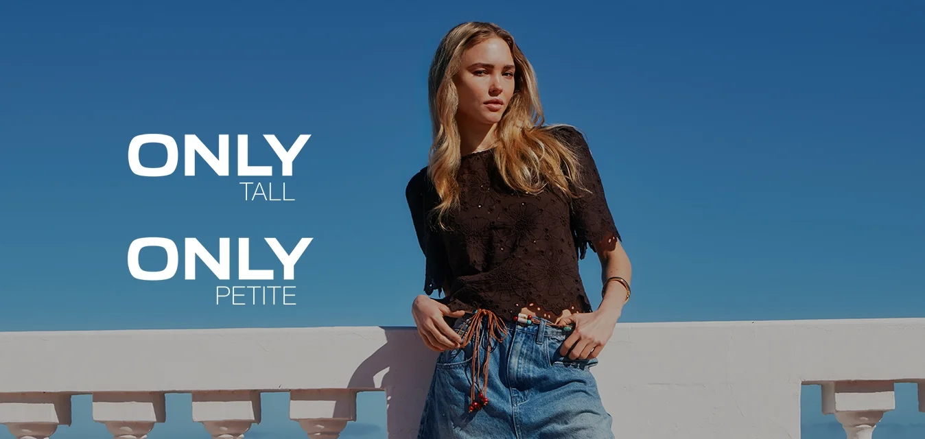 Giovane donna con lunghi capelli biondi che indossa un top nero, pantaloncini di jeans e sandali, si appoggia a una balaustra bianca sotto un cielo azzurro limpido.