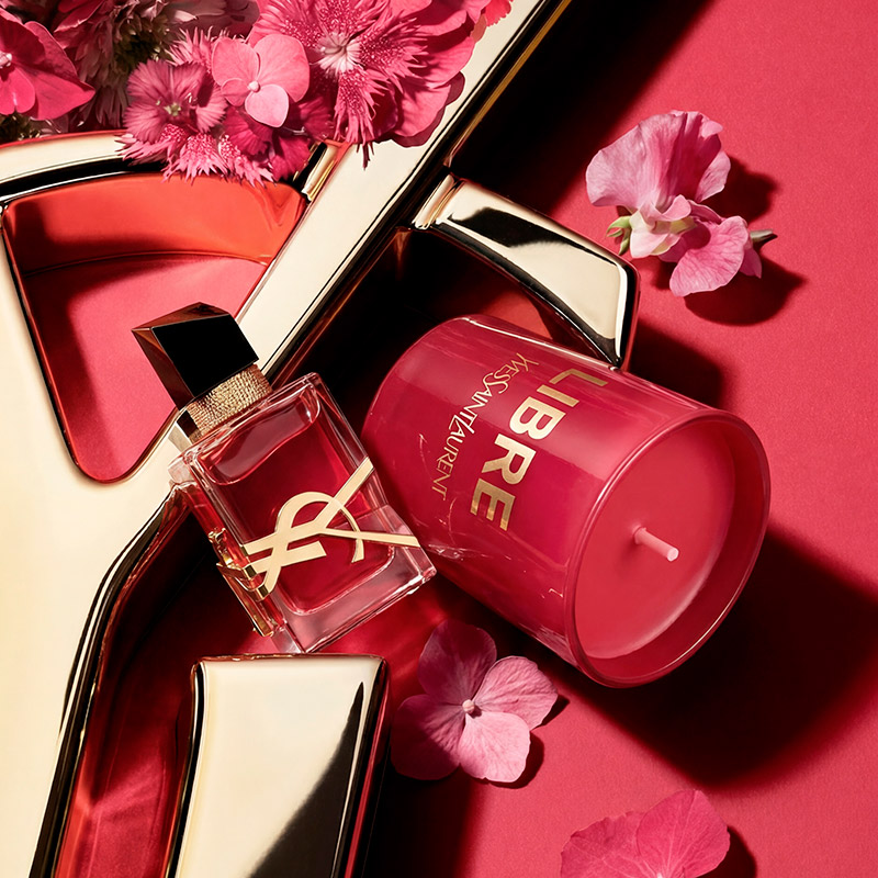 Un flacone di profumo YSL Libre e una candela profumata Libre rosa sono esposti insieme a delicati fiori rosa.