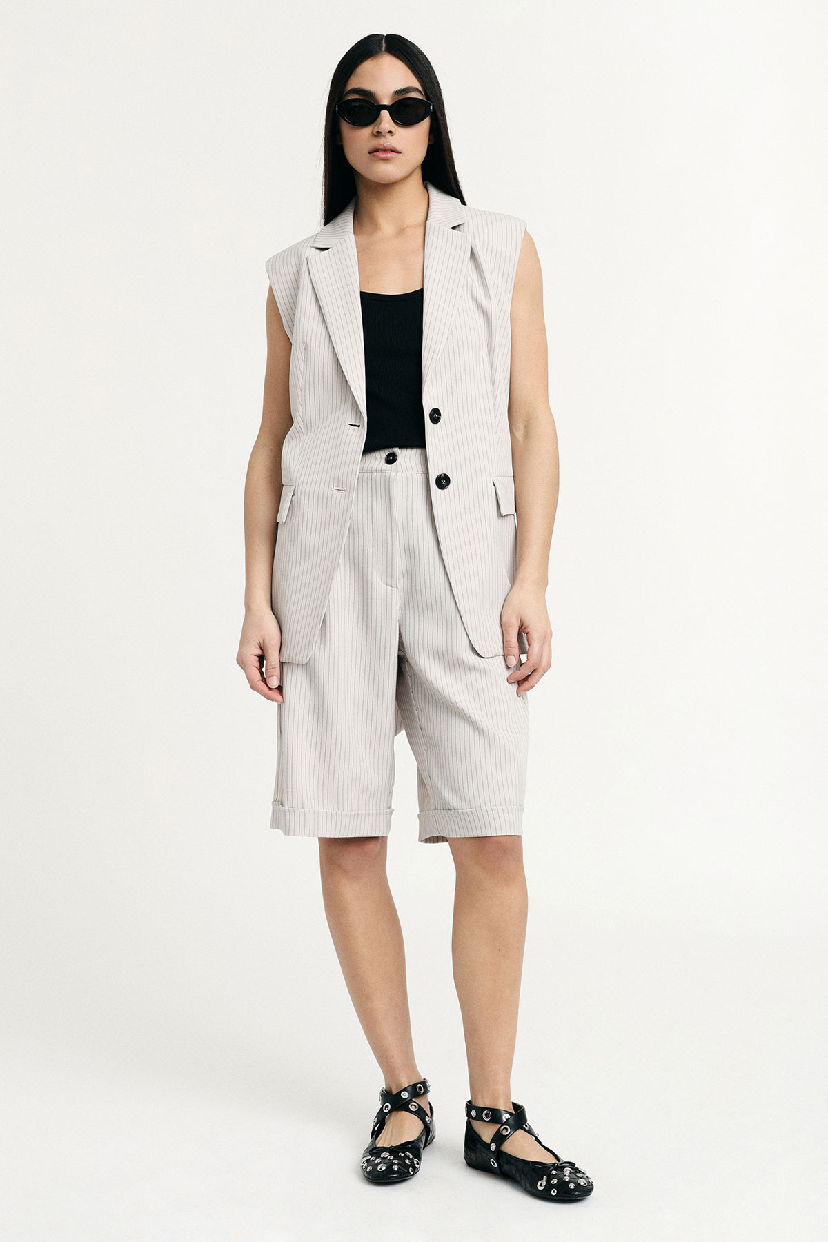 Woman modelling beige pinstripe sleeveless blazer and matching Bermuda shorts with black tank top, cat-eye sunglasses and studded black ballet flats