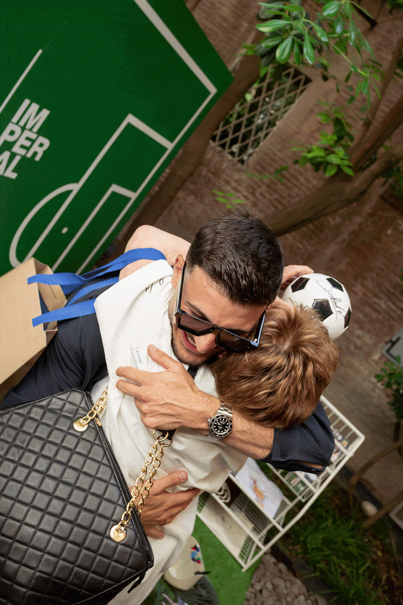 Celebrity at a football event hugging fans and signing autographed jerseys and footballs beside a green 'Imperial' backdrop