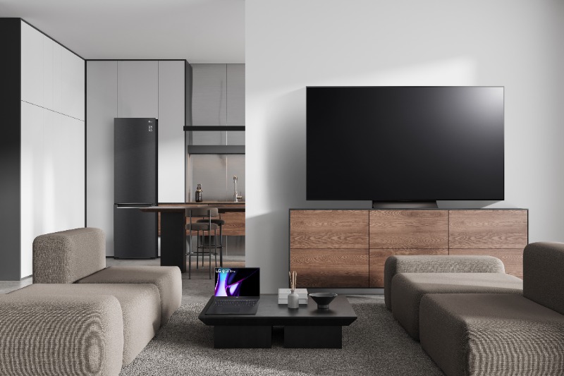 Modern open-plan living room with beige modular sofas, large wall-mounted TV above a wooden media console, black coffee table with laptop and visible kitchen with fridge