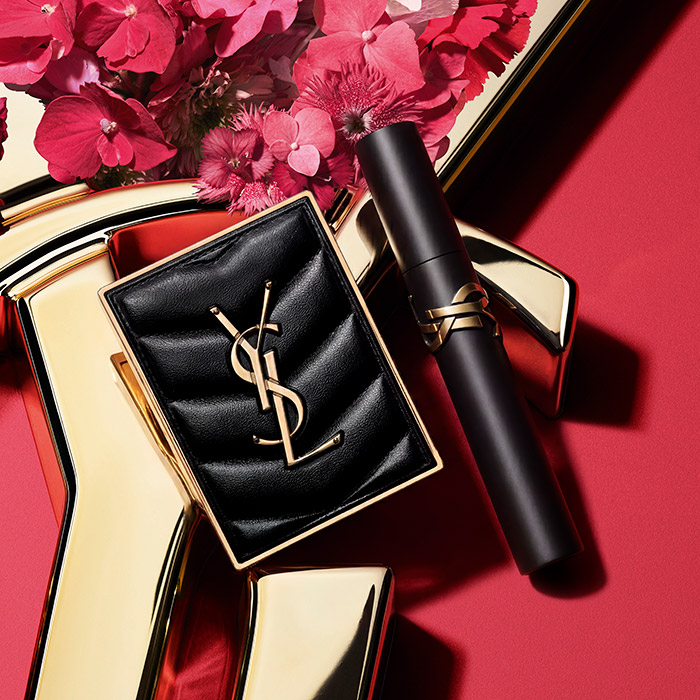 Un compatto trapuntato e un mascara YSL neri esposti con fiori rosa su uno sfondo rosso e oro.