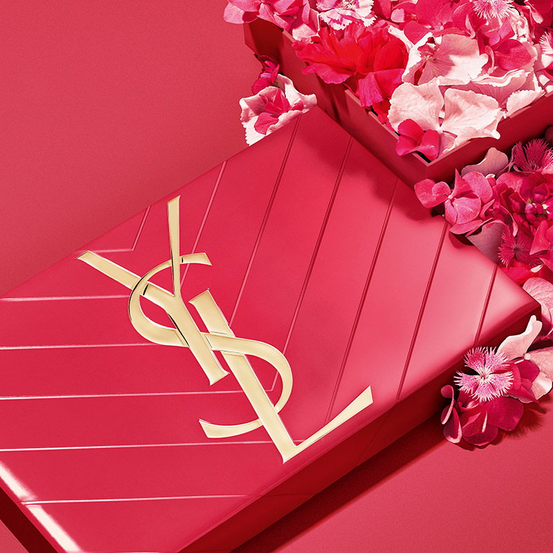 Una confezione regalo YSL rossa trapuntata circondata da fiori rosa.
