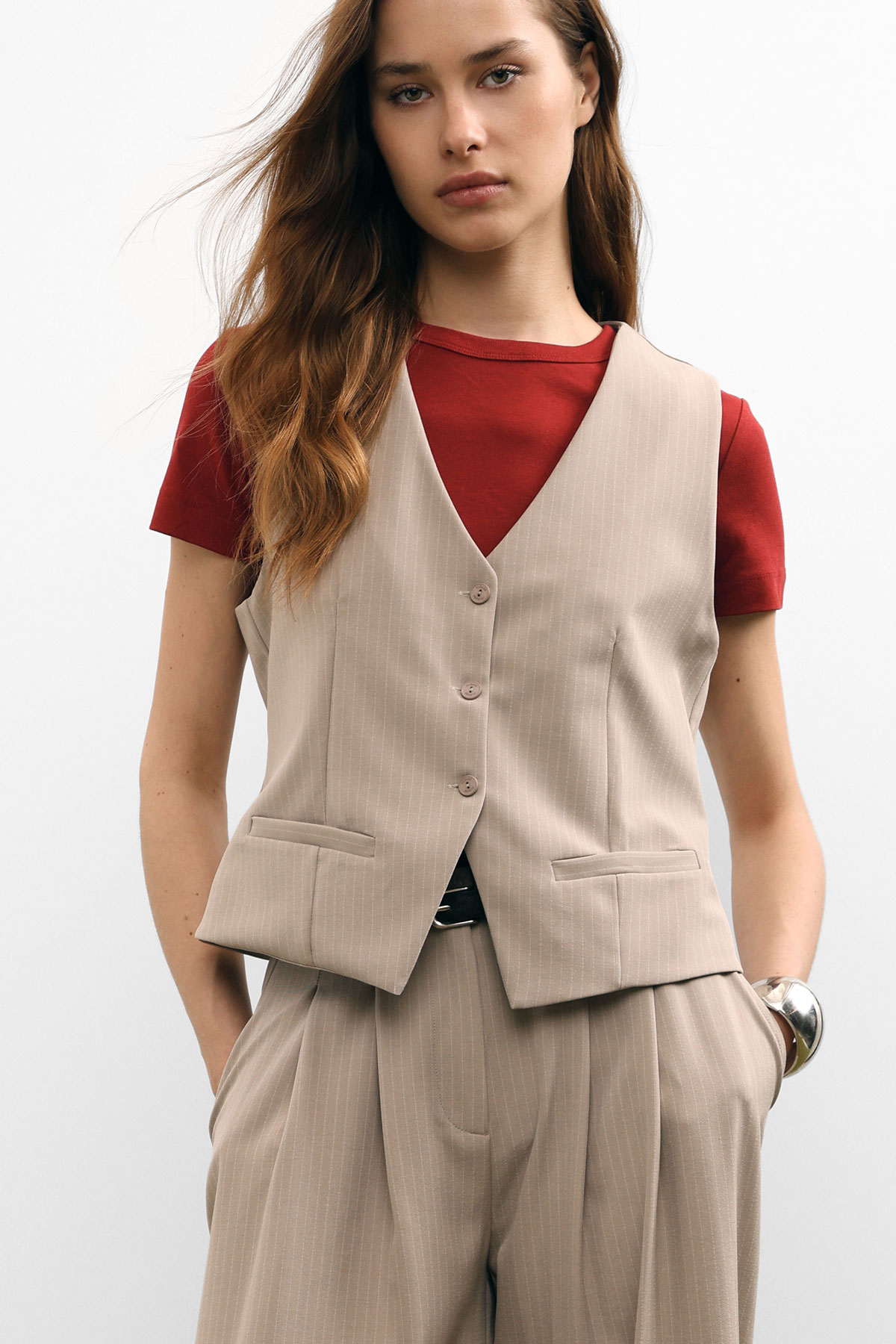 Young woman wearing beige pinstripe waistcoat and matching trousers over a red T-shirt, hands in pockets, neutral studio background