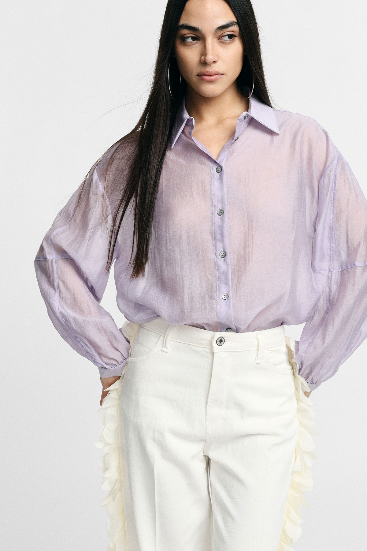 Young woman wearing a sheer lilac button-up blouse and high-waisted white trousers with petal side trim, long dark hair