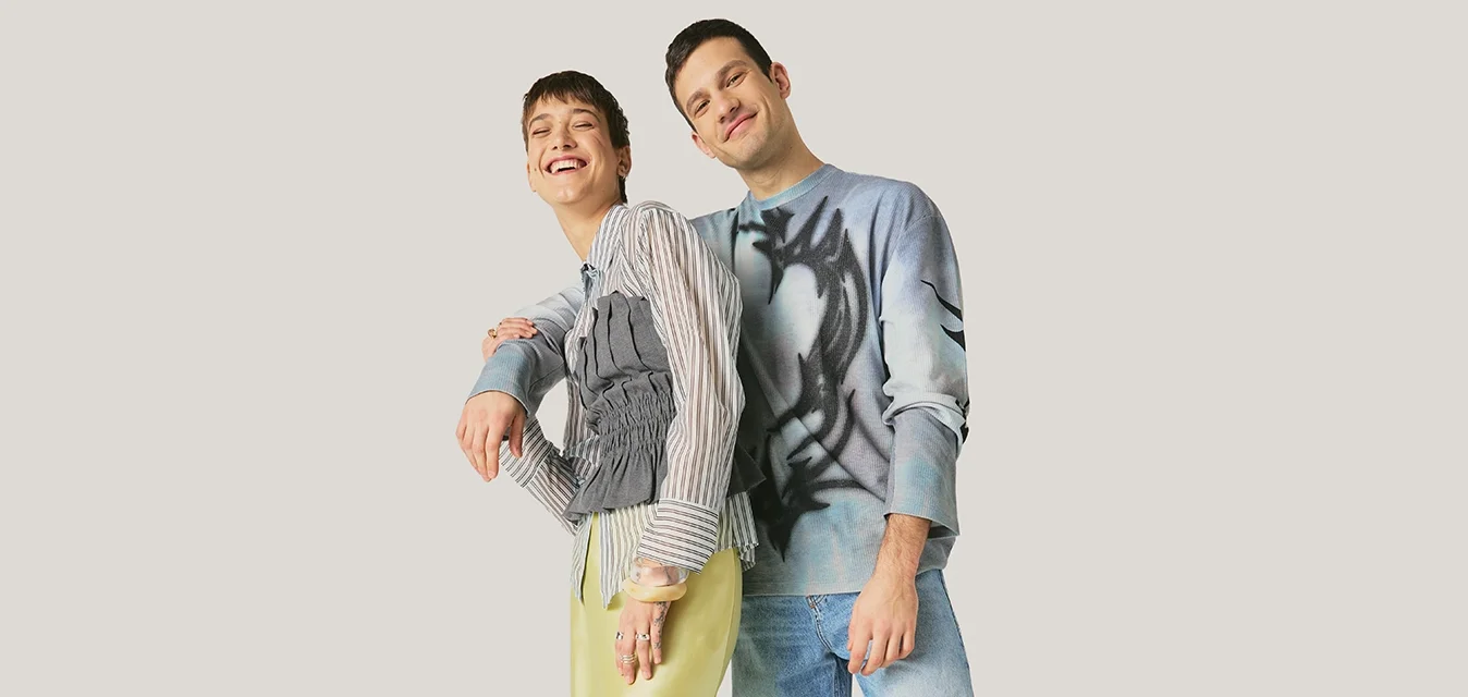 Due giovani adulti stanno vicini l'uno all'altro davanti a uno sfondo semplice, indossano abiti casual moderni con pose rilassate e sorrisi.