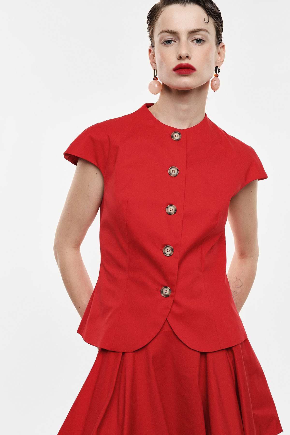Woman wearing tailored red buttoned cap-sleeve blouse and matching pleated skirt with pink drop earrings against a plain background