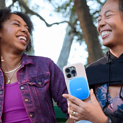 Due donne che ridono, una con in mano uno smartphone con un SSD portatile SanDisk blu collegato.