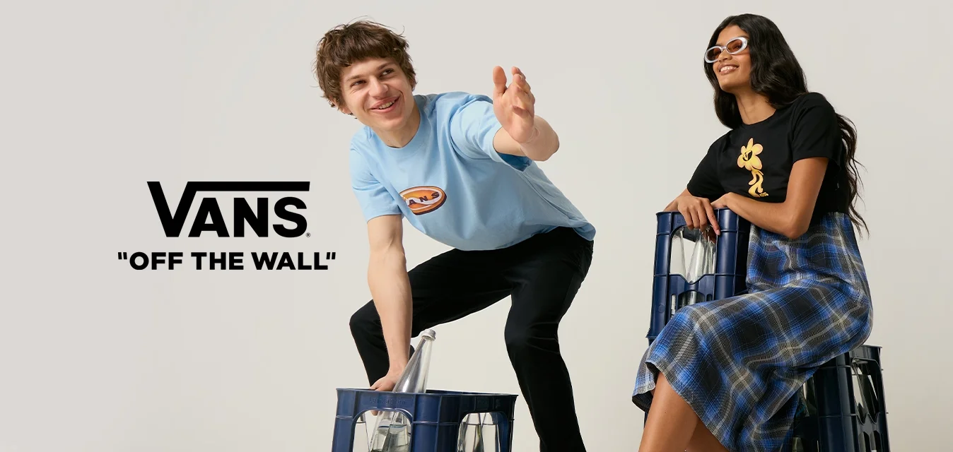 Un giovane uomo si protende in avanti mentre si accovaccia accanto a una cassa, e una giovane donna con occhiali da sole è seduta su una cassa con ruote in uno studio semplice.