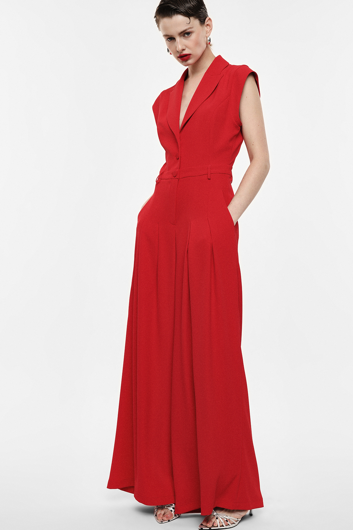 Model wearing sleeveless tailored red jumpsuit with wide palazzo trousers, pockets and waist belt, V-neck against white background