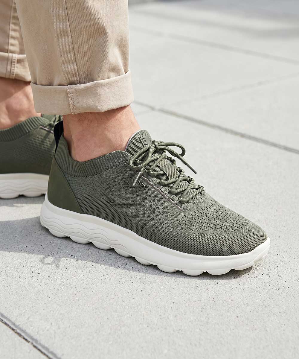 Men's olive-green knit trainers with cushioned white wavy sole and laces, worn on city pavement