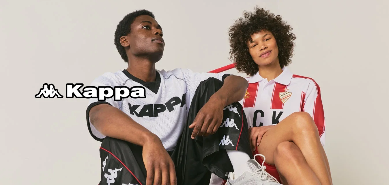 Due persone che indossano abbigliamento sportivo Kappa sono sedute su un carrello a piattaforma rosso contro uno sfondo semplice.