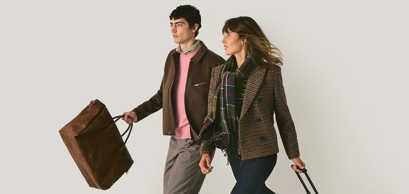 Due persone camminano insieme: una porta una borsa di pelle marrone e l'altra trascina una valigia color sabbia con ruote nere.