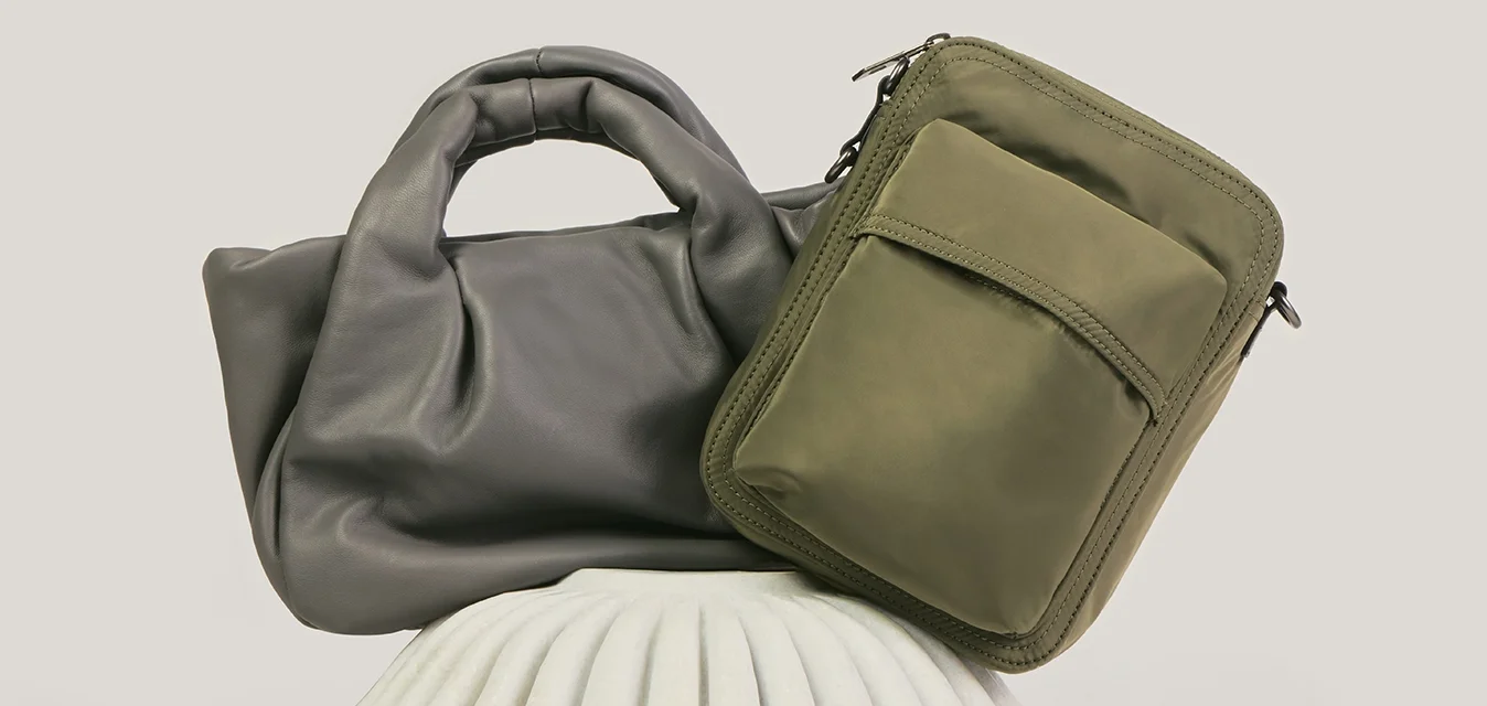 Due borse, una borsa tote in morbida pelle grigia e una tracolla rettangolare verde oliva, esposte su un supporto bianco testurizzato.