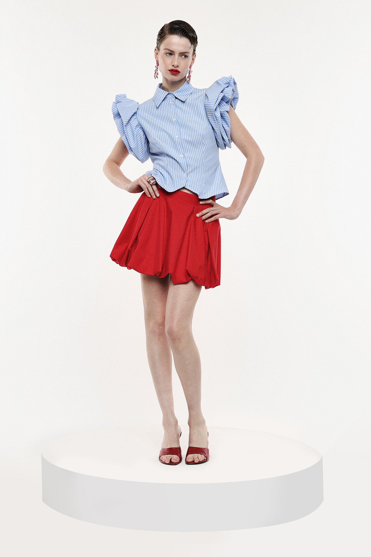 Model on white pedestal wearing blue ruffle-sleeve blouse, red flared mini skirt and red heels