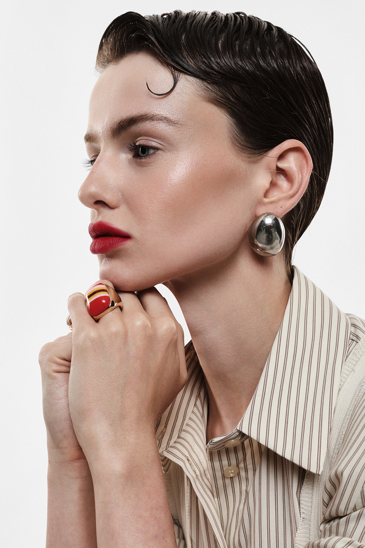Side profile of woman with slicked-back hair, red lipstick, silver earring and striped shirt, hand with red ring by chin