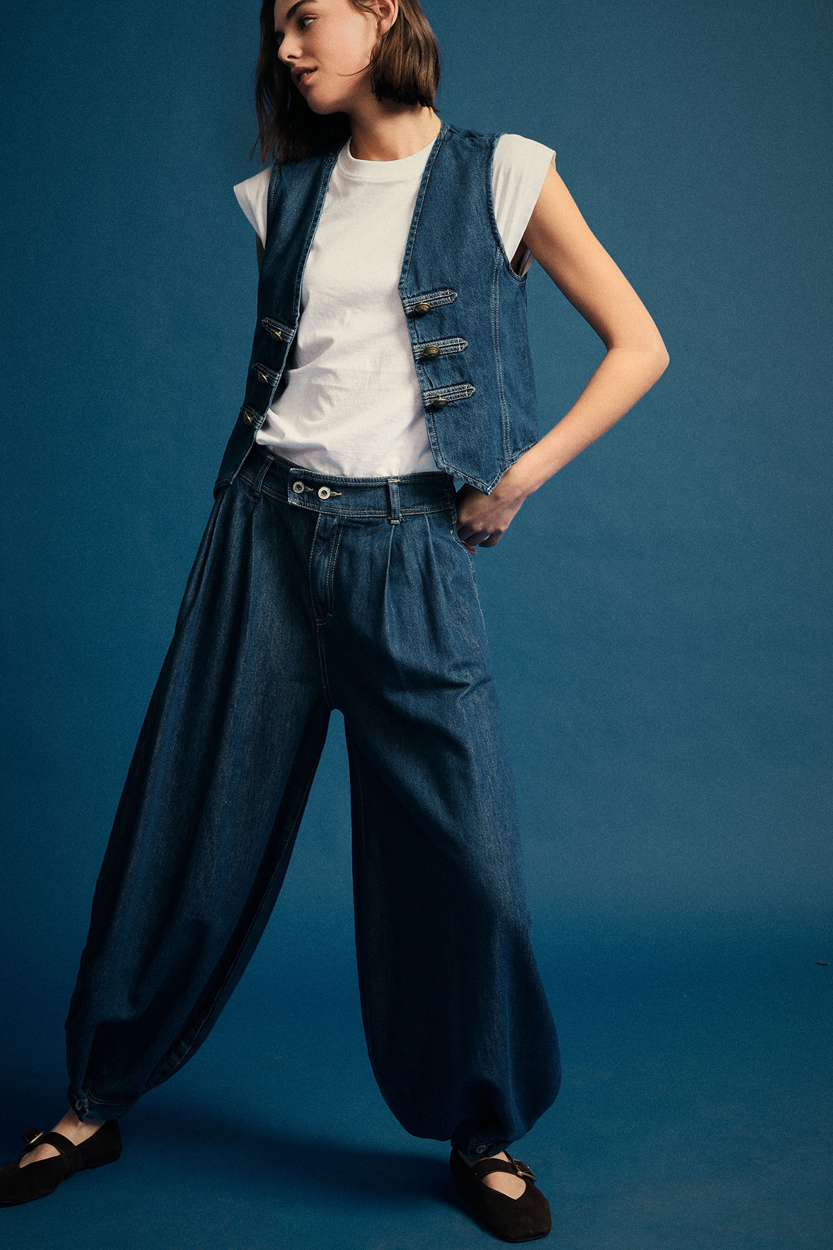Woman wearing white T-shirt, sleeveless denim vest and baggy wide-leg denim trousers with black flats against blue backdrop