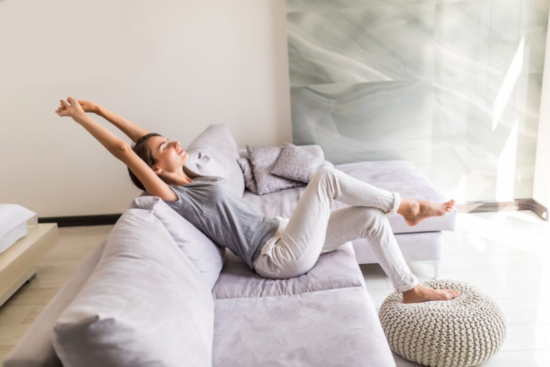 Woman relaxing and stretching on a light-grey sofa in a bright modern living room with cushions and a knitted pouffe