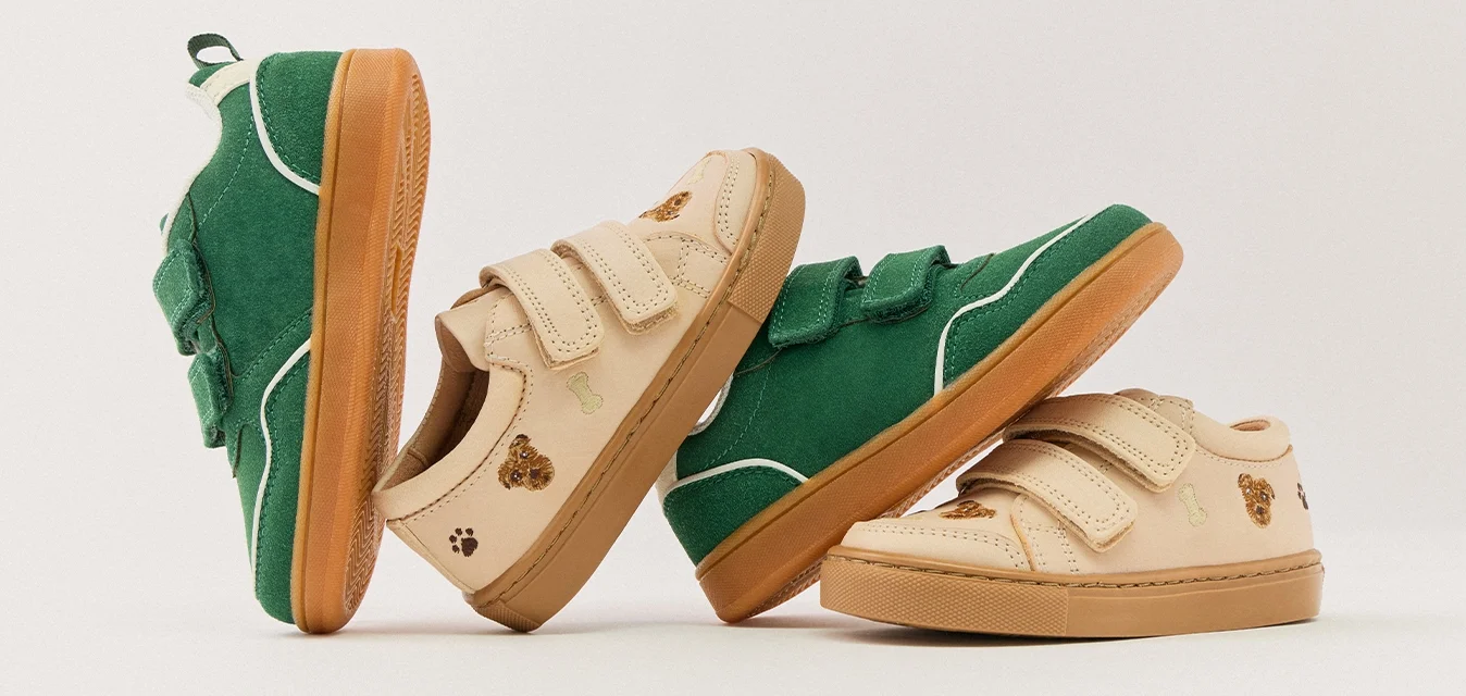 Due paia di scarpe da ginnastica per bambini, una in camoscio verde e l'altra beige con dettagli di orsetto e impronte, disposte su uno sfondo neutro.