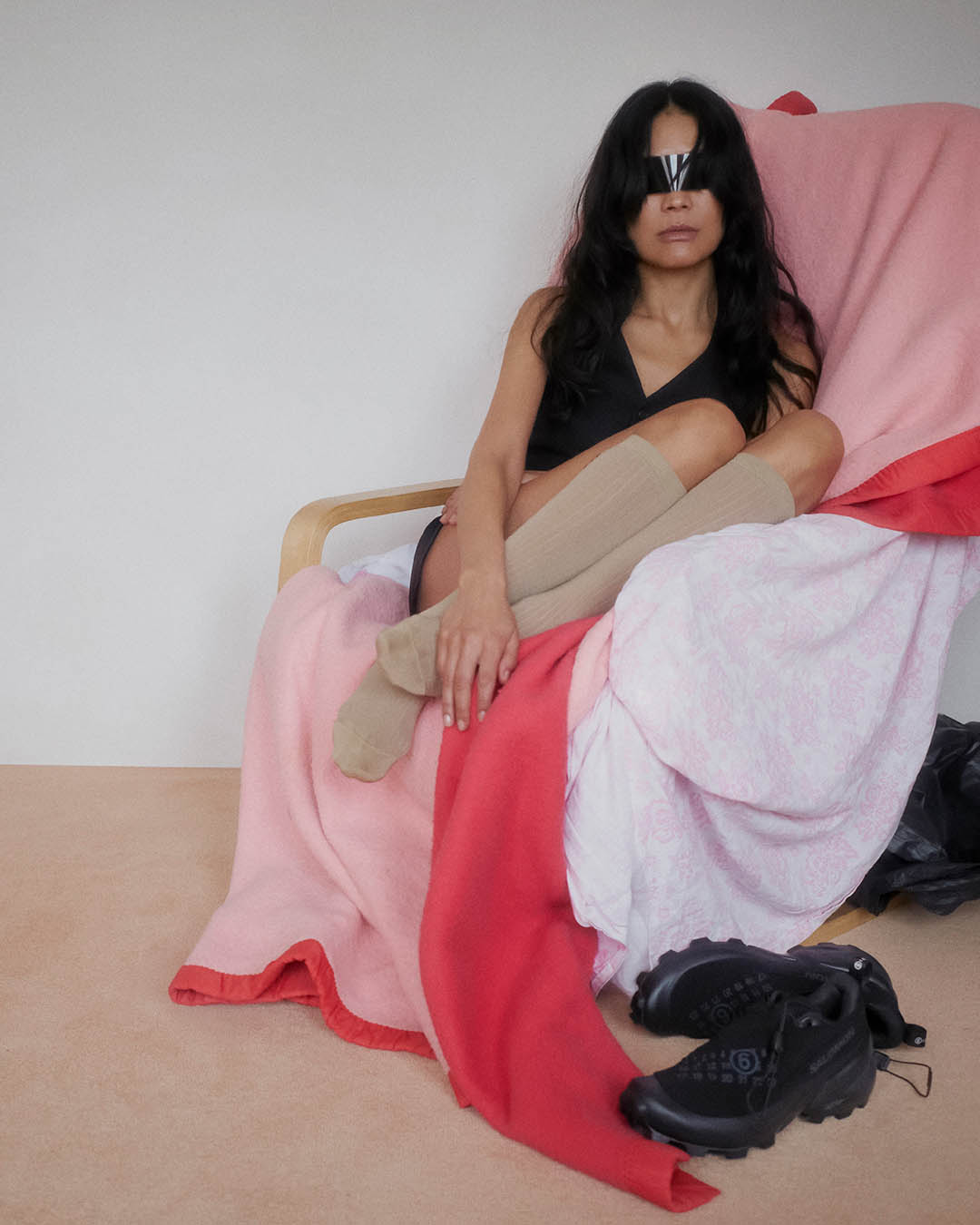 Woman seated on armchair draped with pink and red blankets, wearing a black sleeveless top, beige knee socks and reflective eye-covering, black shoes on floor