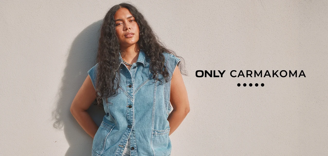 Donna con capelli lunghi e ricci che indossa un gilet di jeans senza maniche e pantaloni azzurri, in piedi contro un muro semplice di colore chiaro.