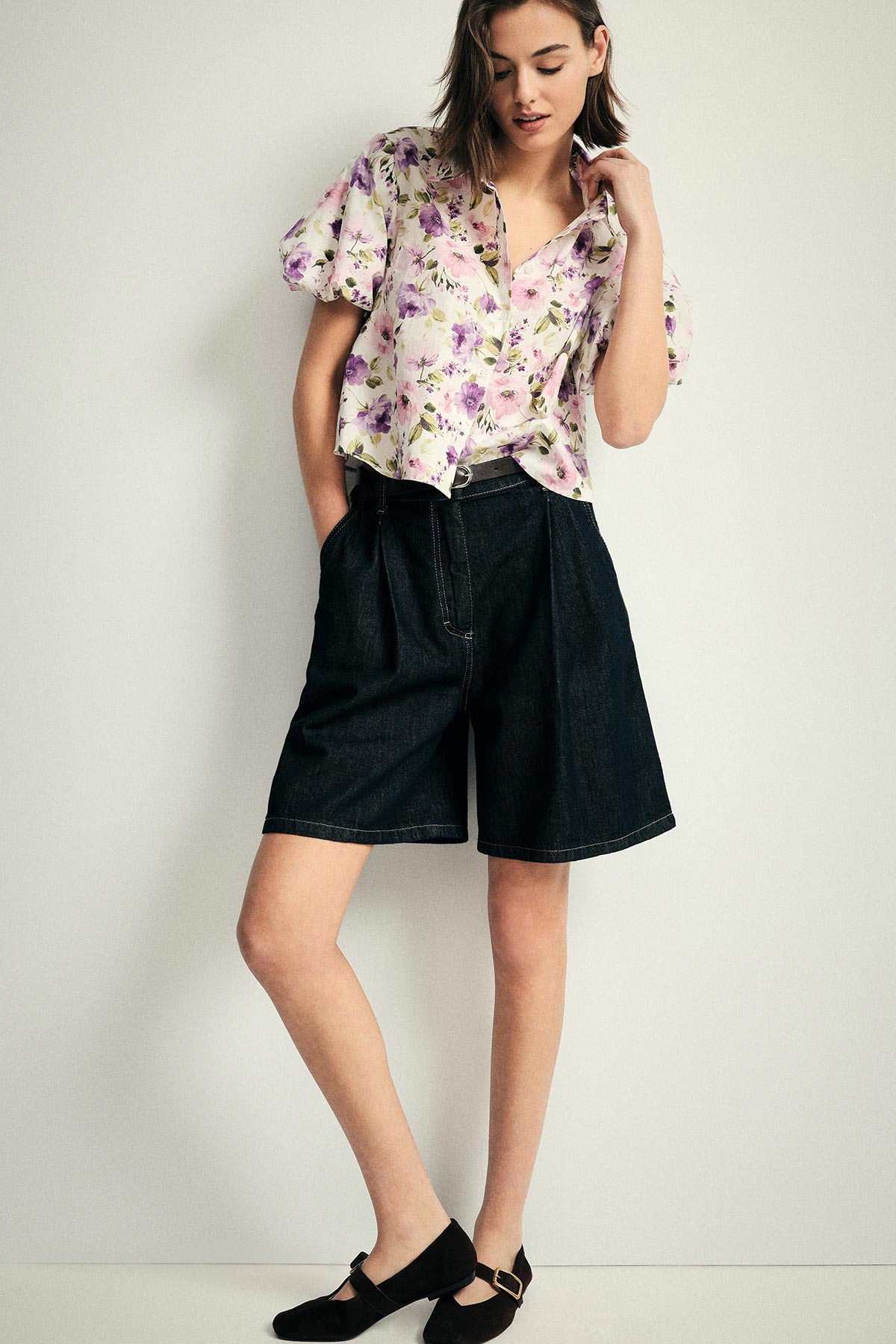Woman standing against a plain background wearing a pastel floral blouse, high‑waisted dark denim knee‑length shorts and black Mary Jane shoes