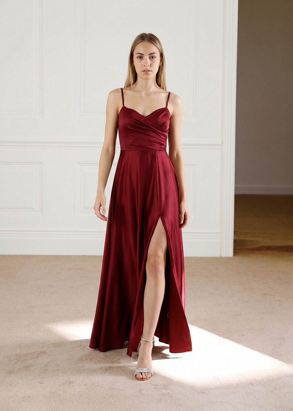 Woman in burgundy satin evening gown with spaghetti straps and thigh-high slit walking in bright room wearing silver heels