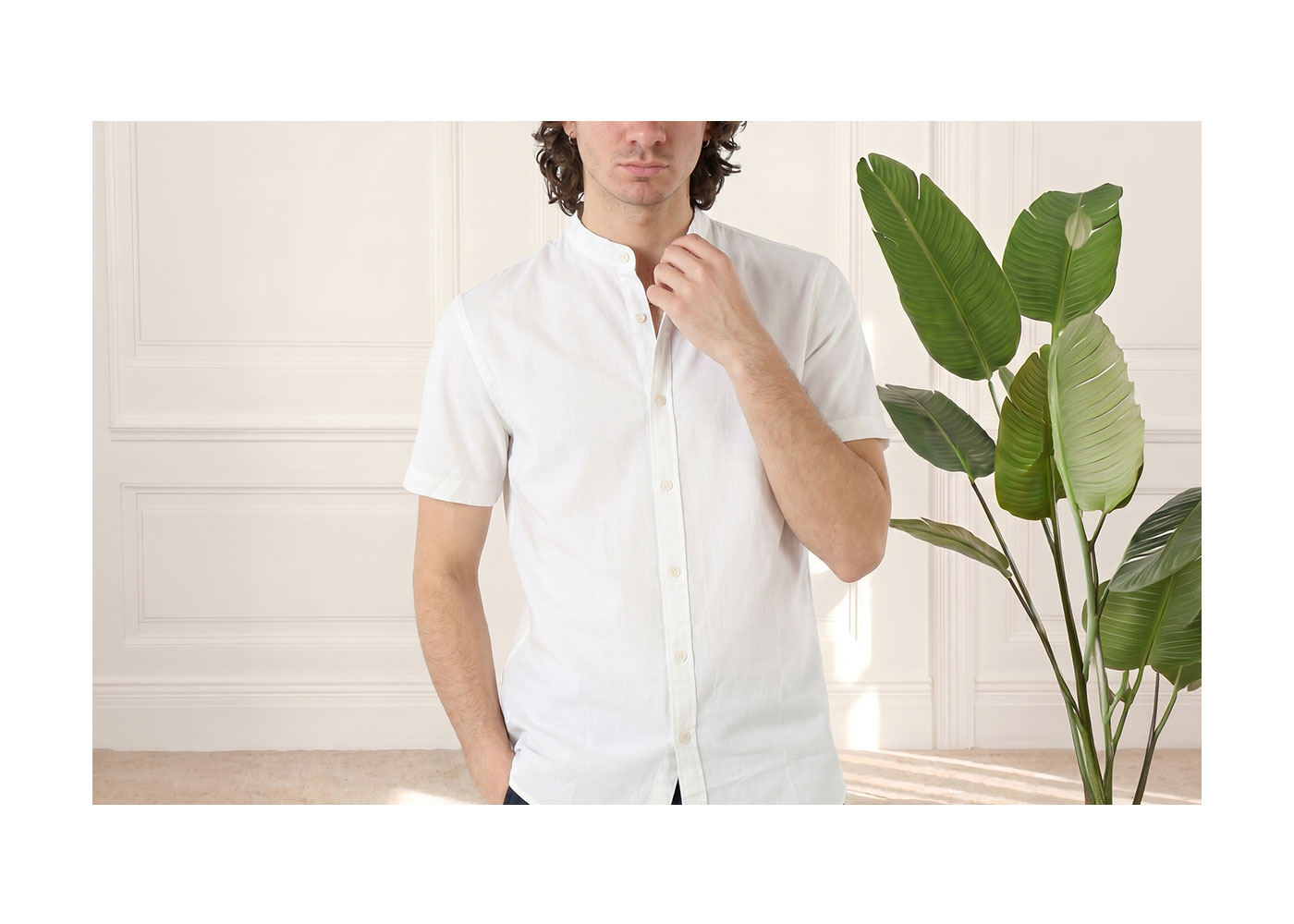 Man wearing white short-sleeved linen shirt with mandarin collar, adjusting collar beside large green houseplant in bright room
