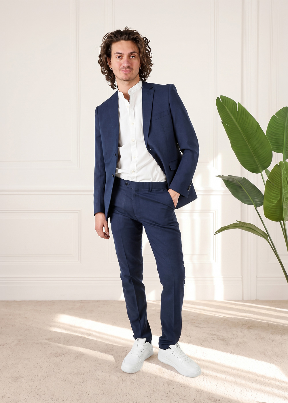 Man modelling a navy suit with white shirt and white trainers, standing in a bright room beside a large potted plant