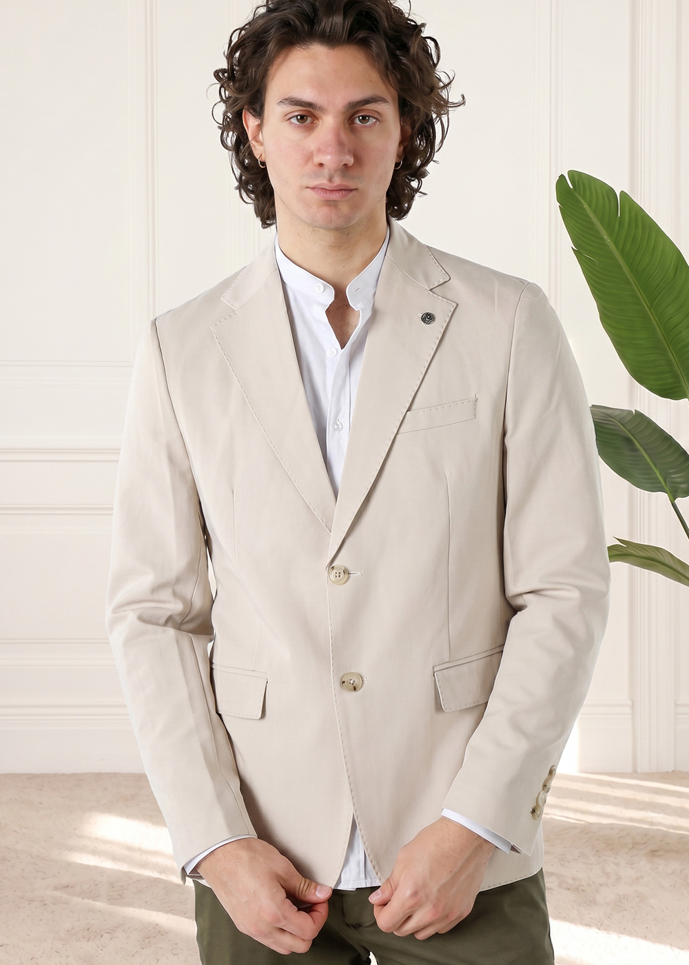 Man wearing a beige blazer and white shirt in a smart-casual outfit, standing indoors with a green plant in the background