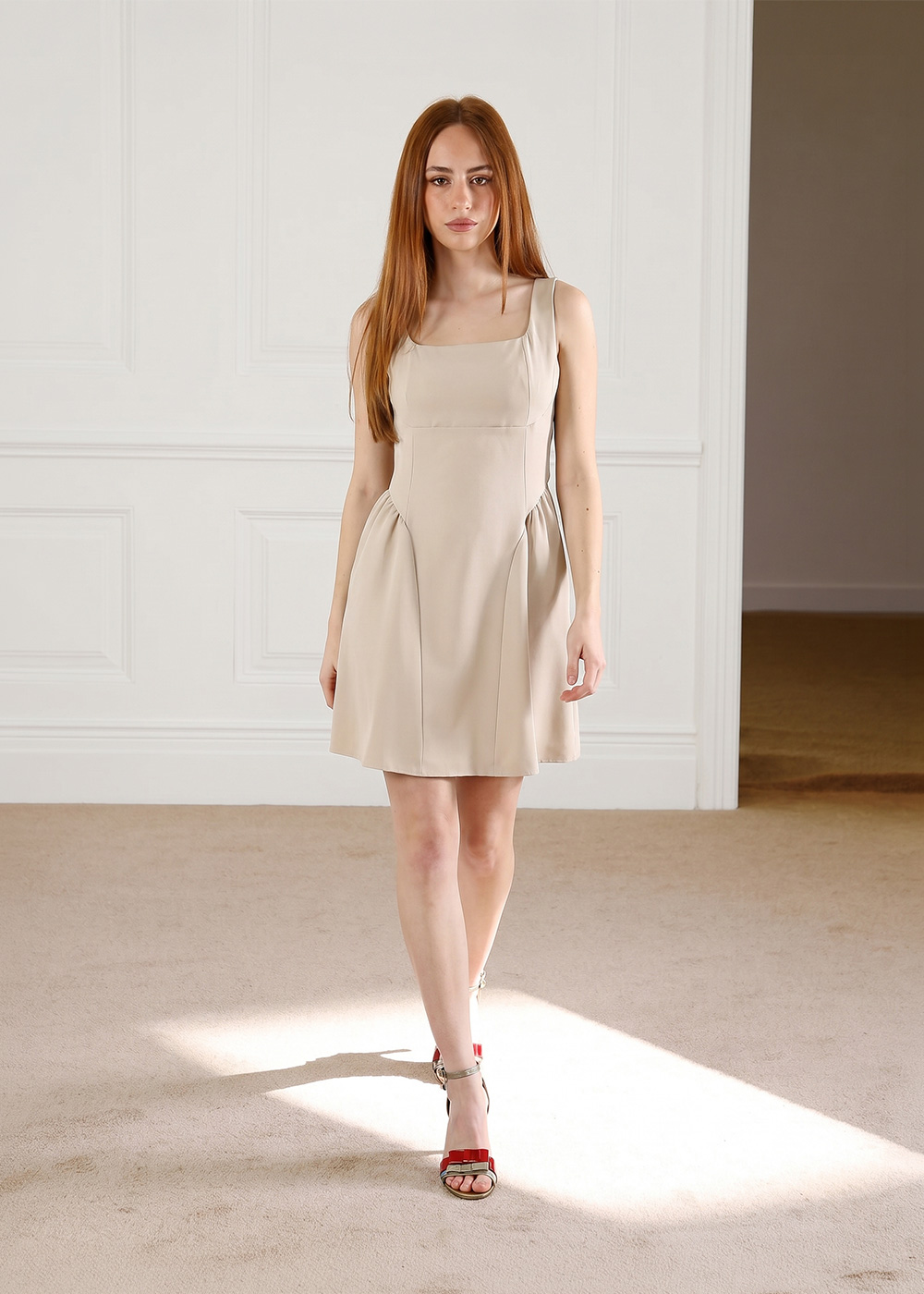 Woman in sleeveless beige mini dress walking indoors on a carpeted floor wearing red heeled sandals